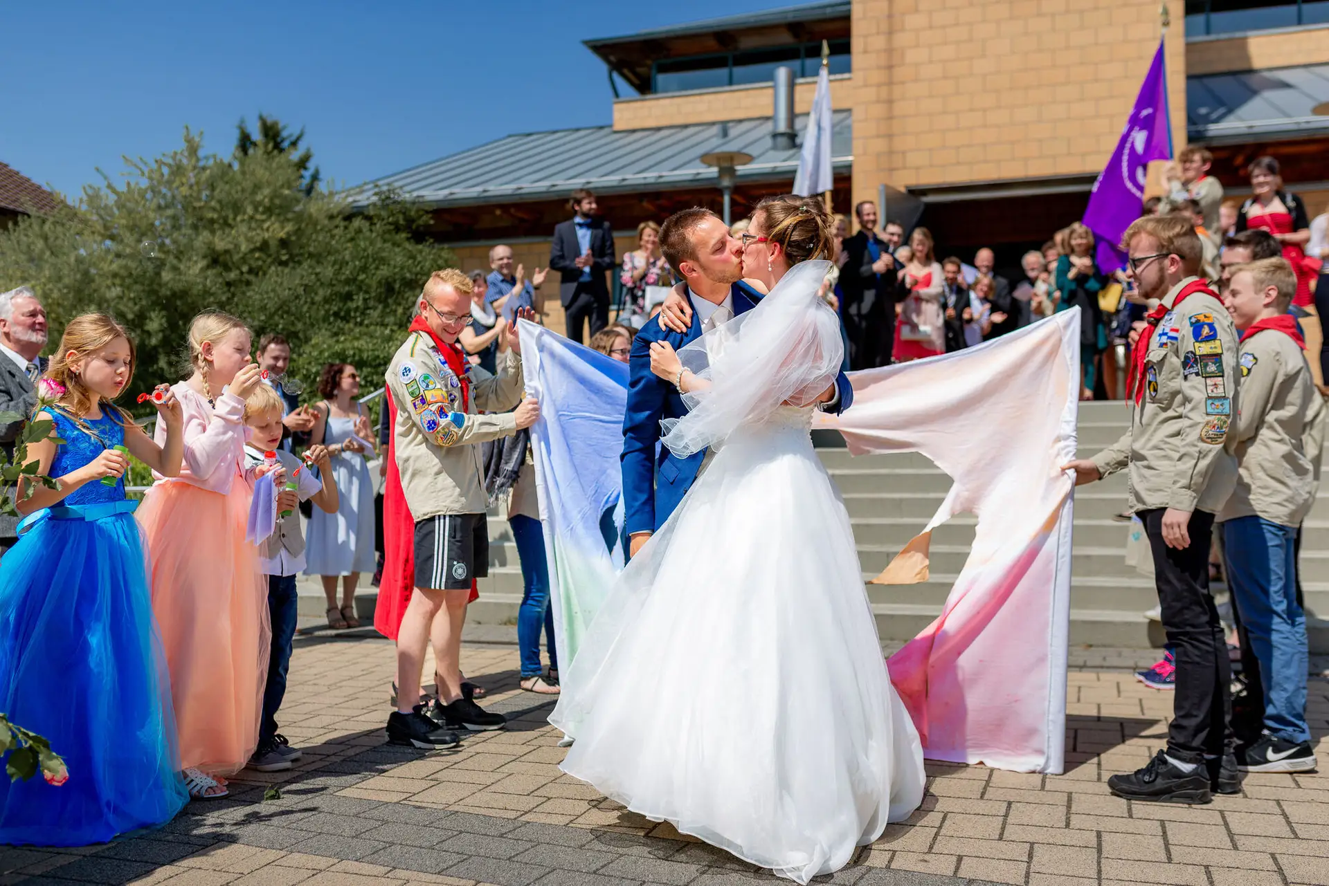 Braut küsst Bräutigam freudig vor dem Gebäude, während Gäste mit bunten Fahnen jubeln