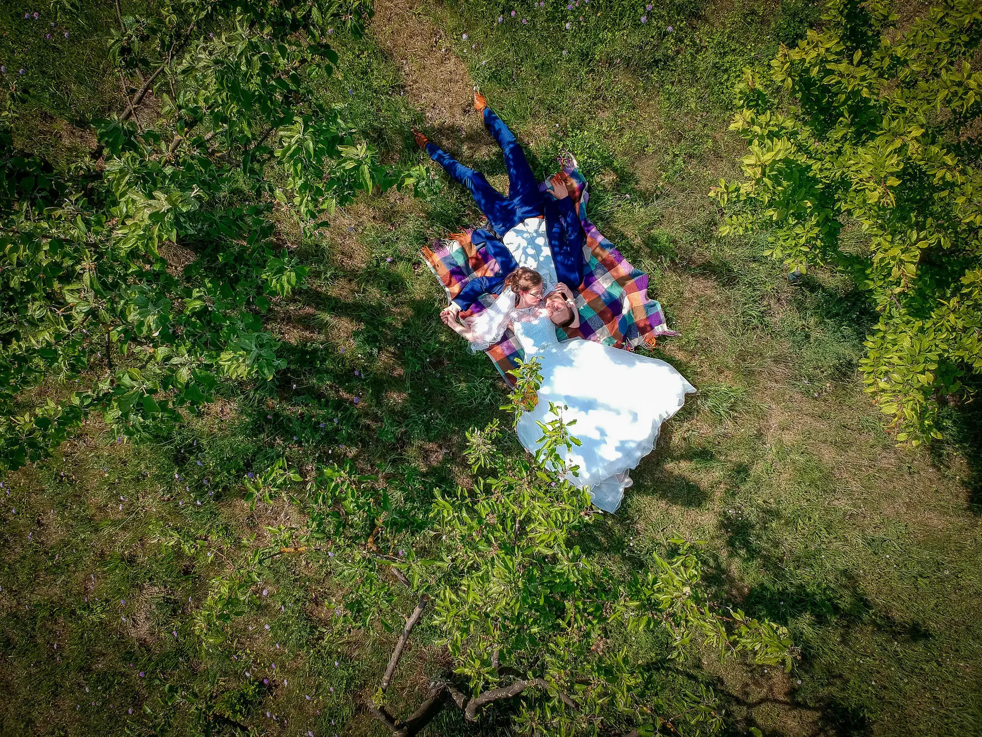 Hochzeitspaar liegt entspannt auf einer Decke im Grünen, ruht sich unter schattigen Bäumen aus