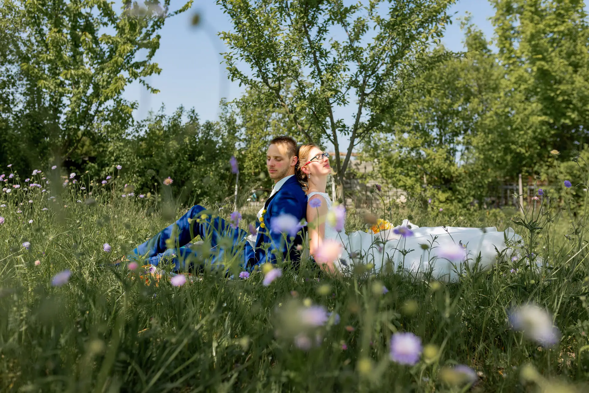 Hochzeitspaar sitzt auf Wiese mit Lavendelblüten im Hintergrund und blickt gemeinsam in die Ferne