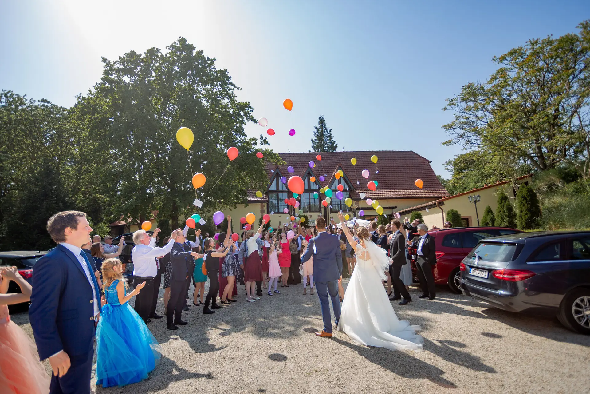 Brautpaar stößt lachend mit bunten Luftballons an, Gäste jubeln rundum beim Sektempfang im Garten