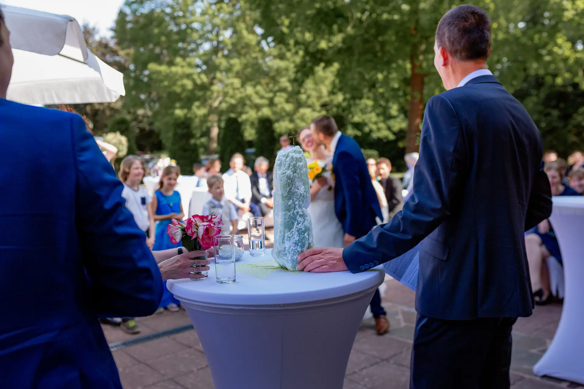 Trauzeugenleiter führt das Brautpaar beim Hochzeitsritual vor einem weißen Blumentisch mit Grünpflanzentuch