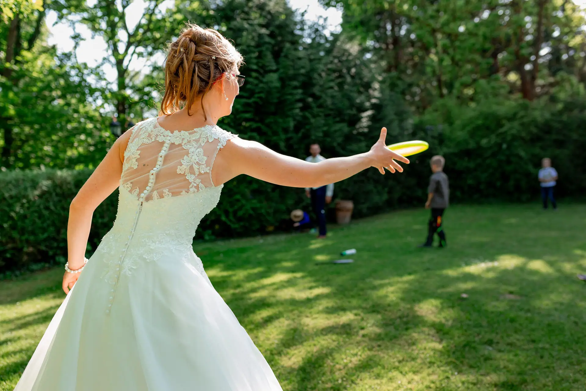 Braut wirft ausgelassen einen Frisbee in den grünen Hochzeitsgarten im weißen floralen Kleid