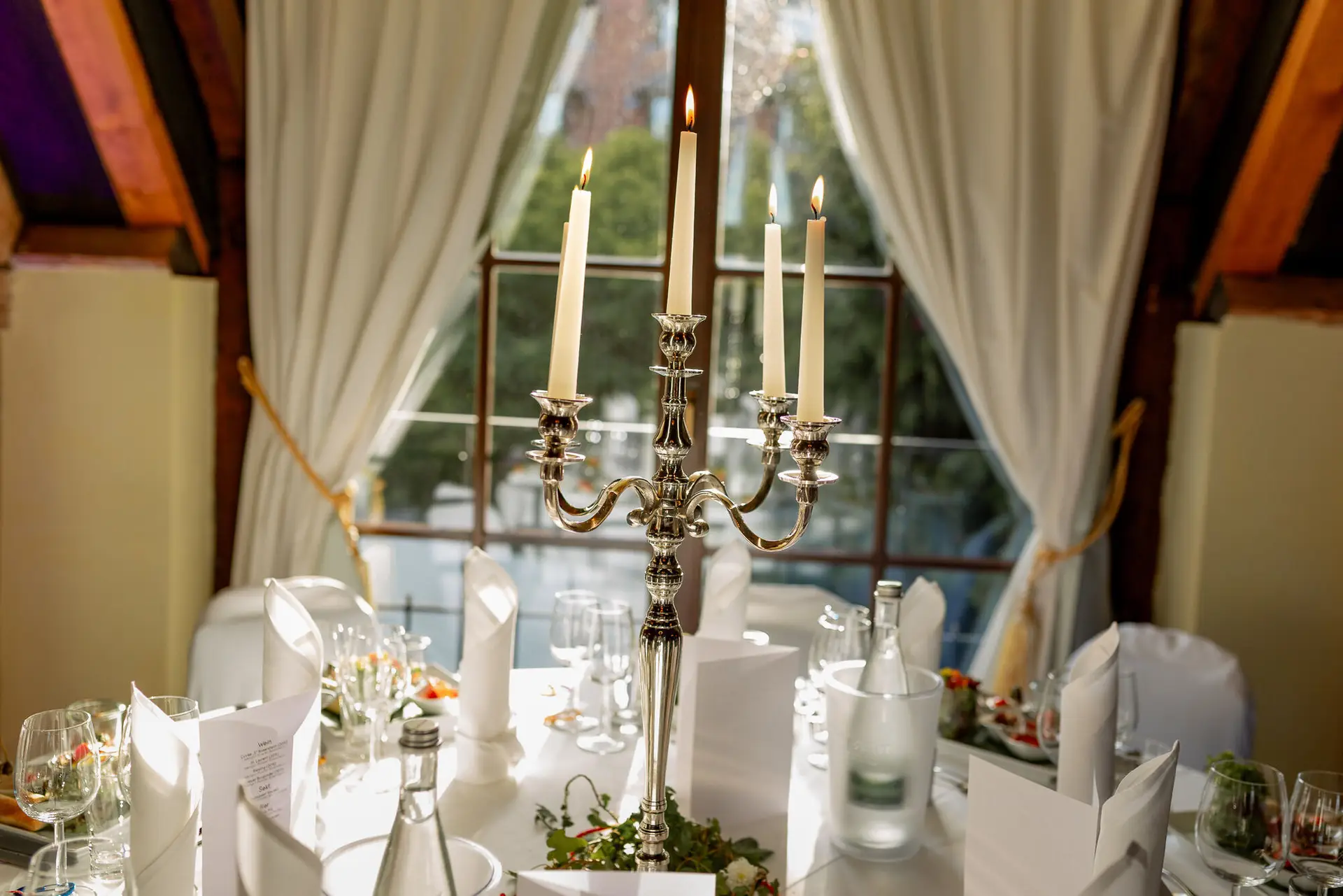 Silberne Kerzenleuchter mit Kerzen auf gedecktem Hochzeitstisch mit Menükarte vor dem Saalfenster