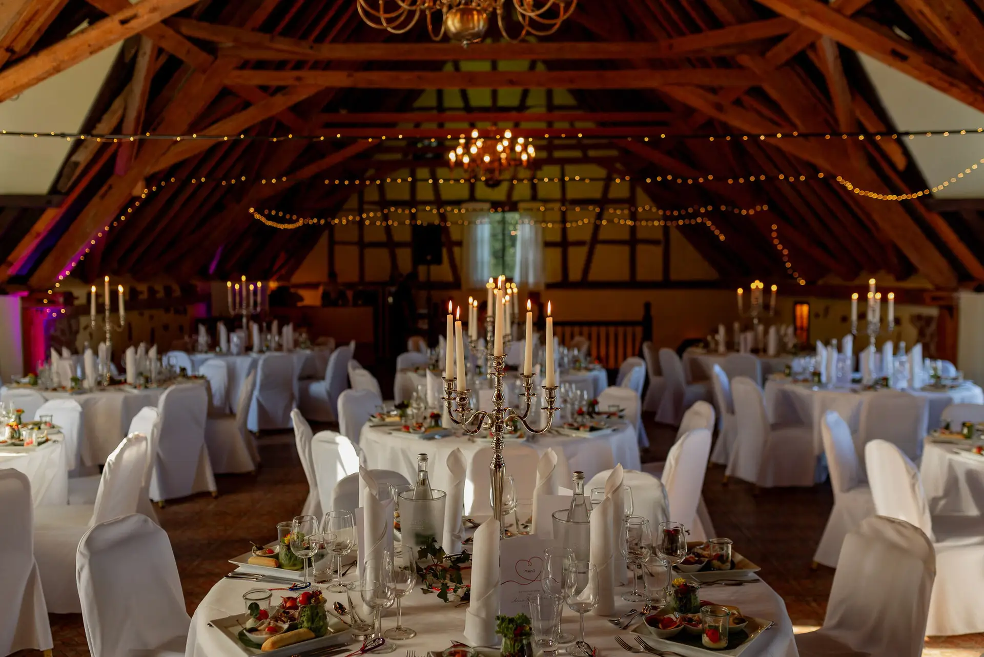 Hochzeitsdinner in strohgedecktem Saal mit Kerzen, weißen Tischdecken und Gästen an langen Tafeln