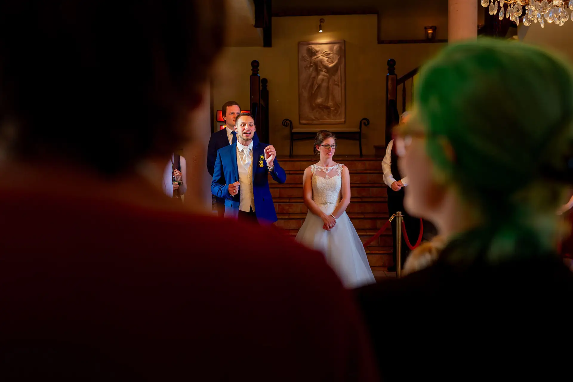 Bräutigam steht elegant in historischem Saal mit Treppe und Kristalllüstern, Braut tritt im Hintergrund ein