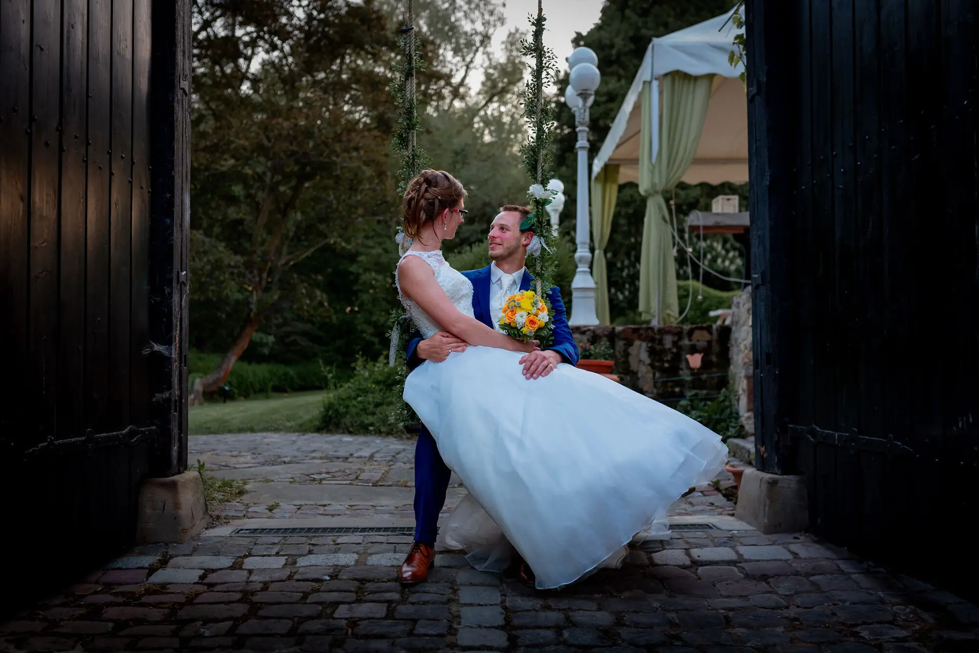 Brautpaar sitzt eng umschlungen an einem Holzzaun unter grünem Pavillon mit gelben Blüten
