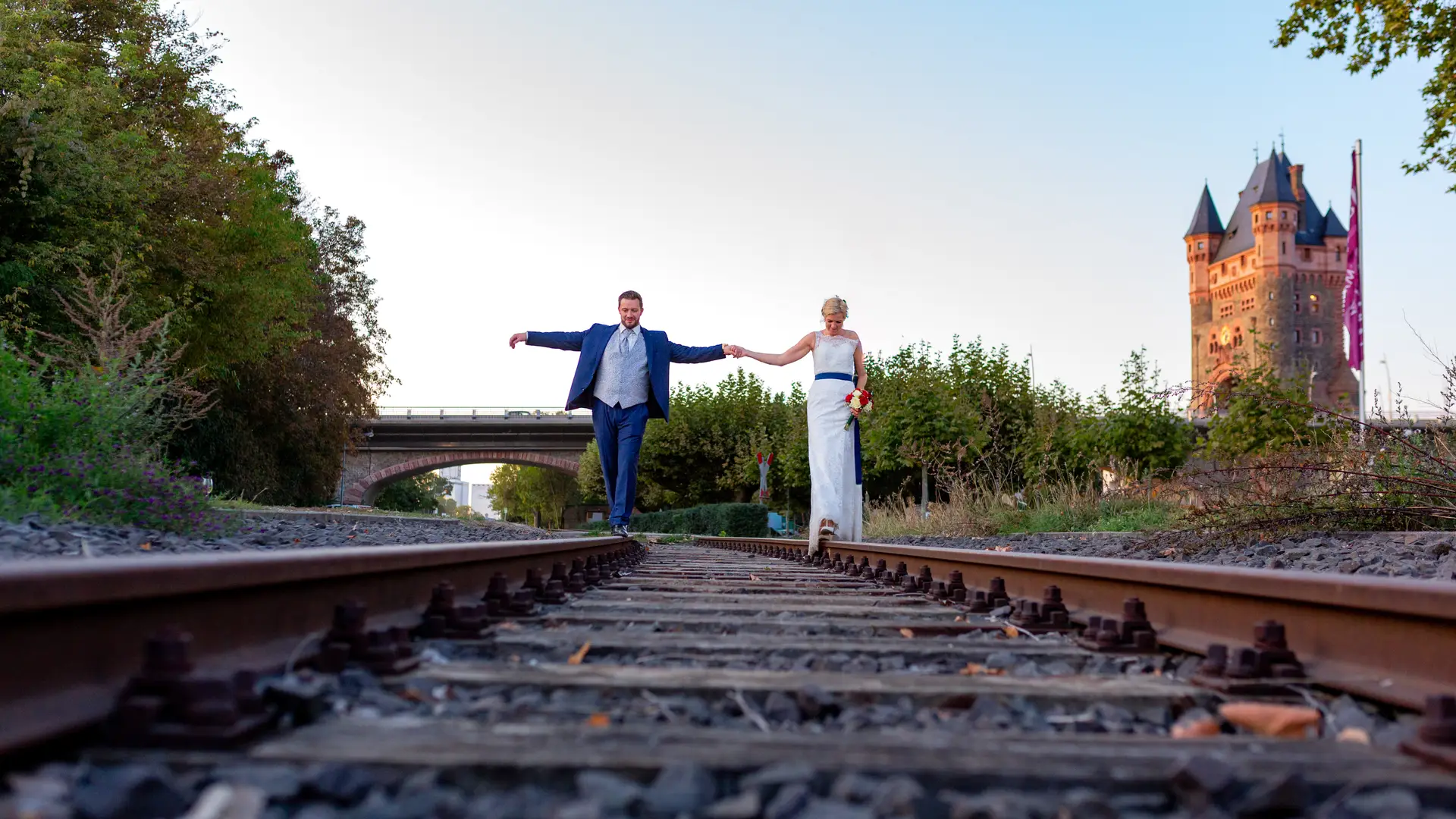 Brautpaar balanciert Hand in Hand auf Gleisen in Worms, Nibelungenturm im Hintergrund