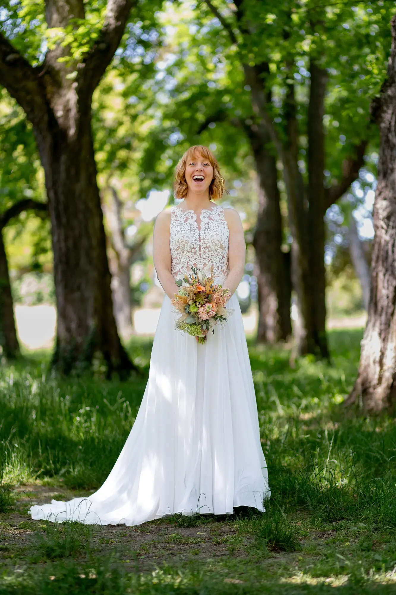 Braut in weißem Spitzenkleid mit Boho-Blumenstrauß lacht strahlend im Waldbereich des Schlossgartens