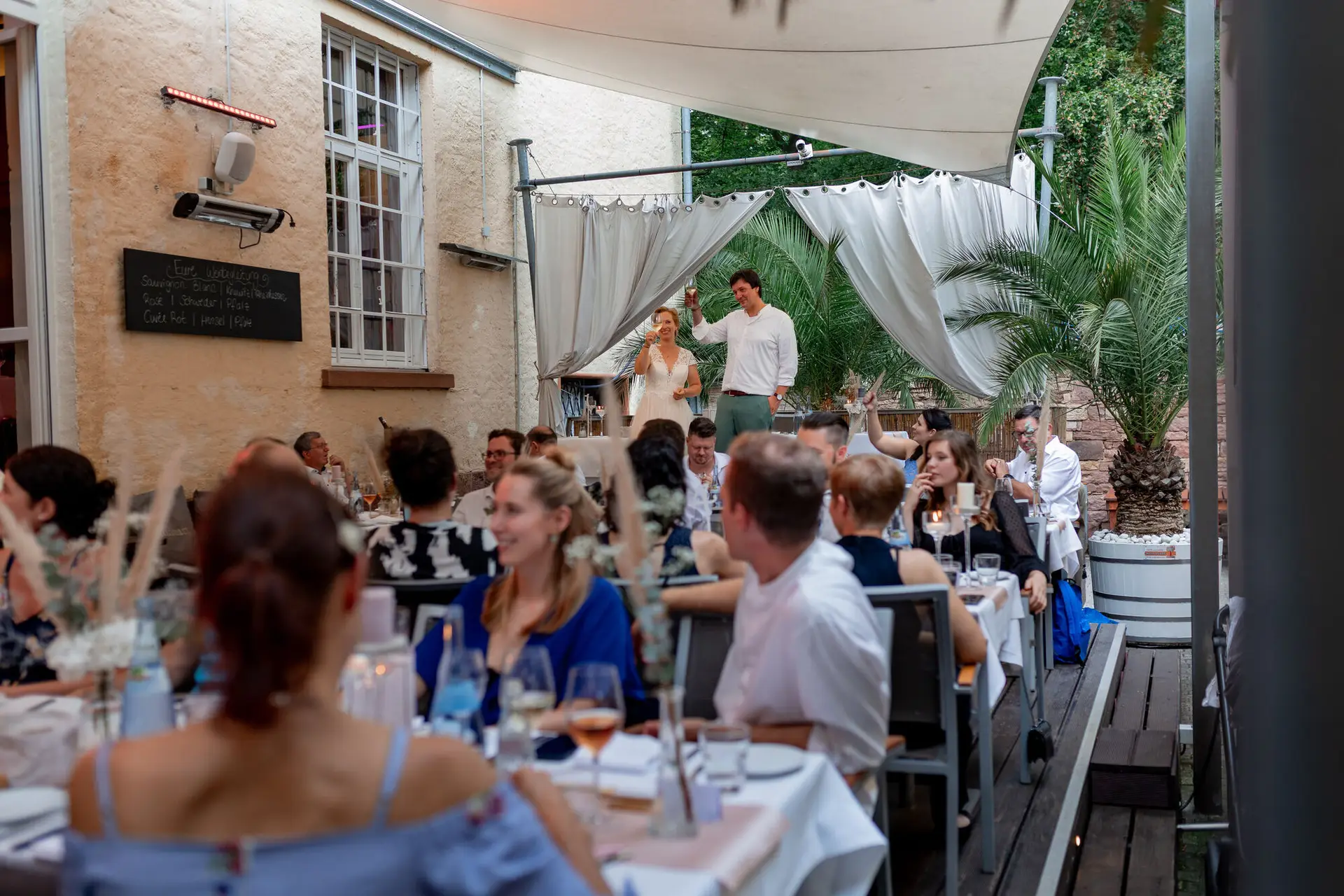 Hochzeitsgesellschaft beim Abendessen auf der Terrasse, Brautpaar erhebt die Gläser zu einem Toast