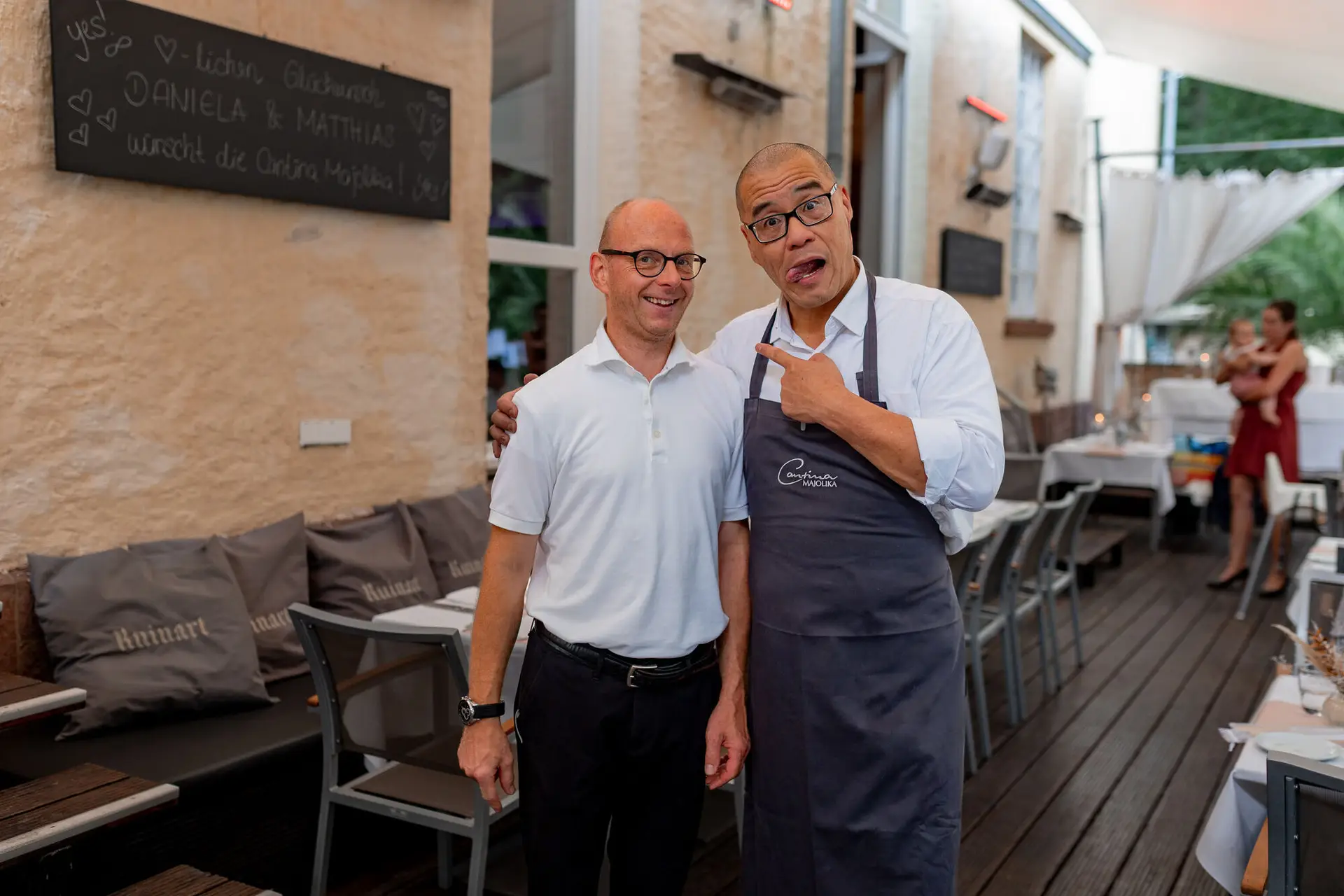 Gast und Koch posieren zusammen vor der Tafel mit Daniela & Matthias auf der Terrasse der Cantina Majolika