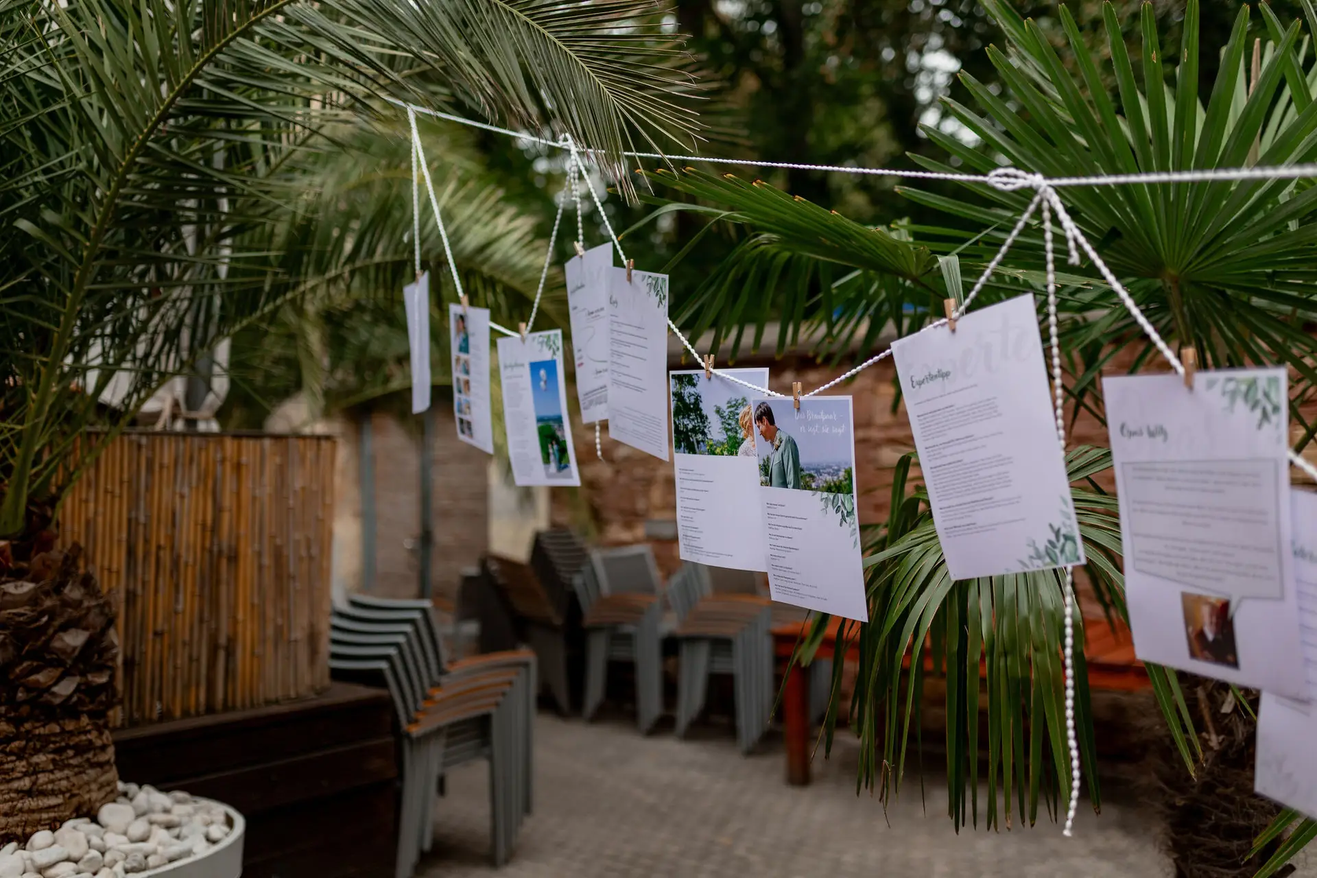 Hochzeitskarten hängen an einer Leine zwischen Palmenblättern über Steinmauer mit Sitzgelegenheiten
