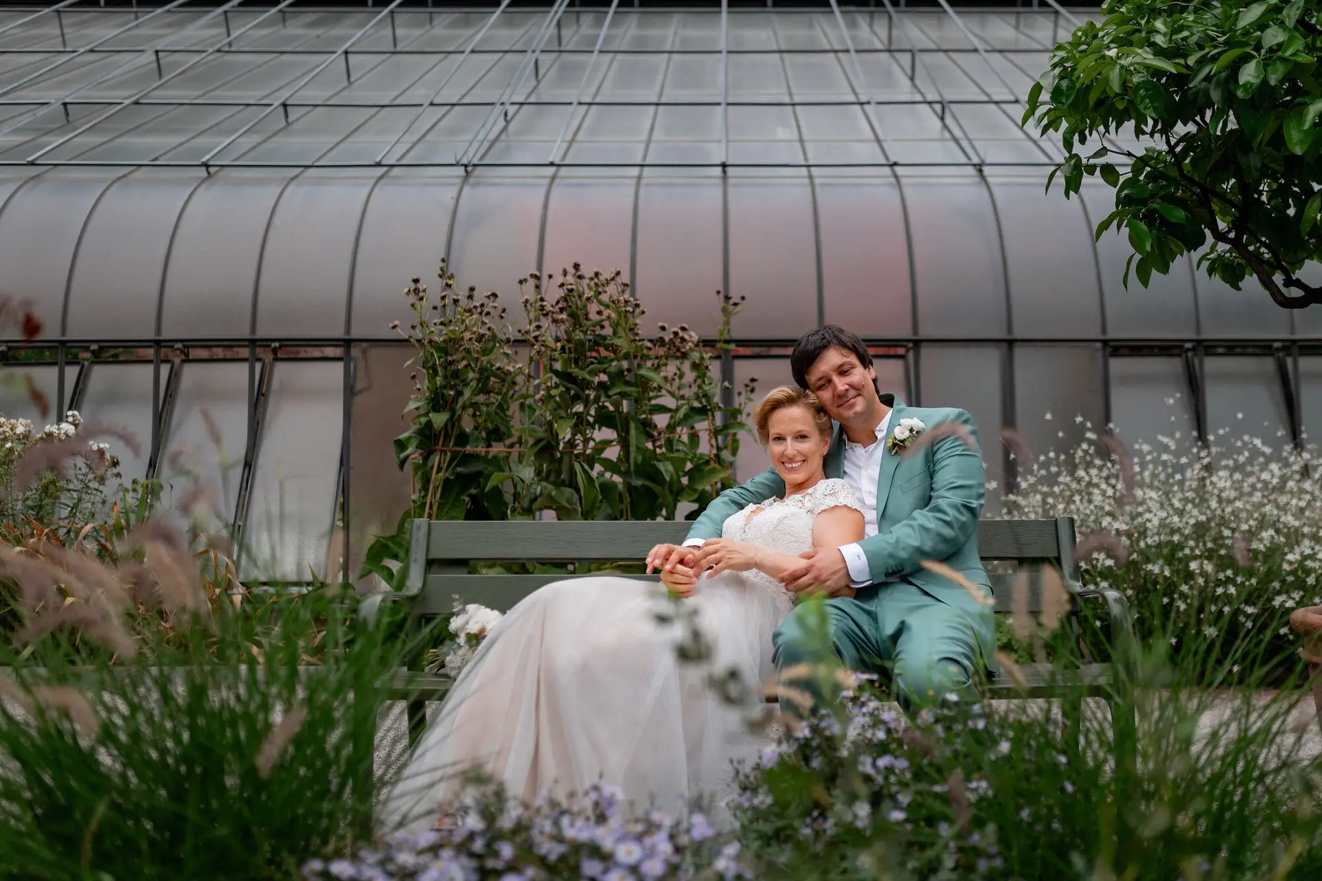 Brautpaar sitzt eng aneinandergeschmiegt auf einer Bank im Gewächshaus, umgeben von üppig blühenden Pflanzen