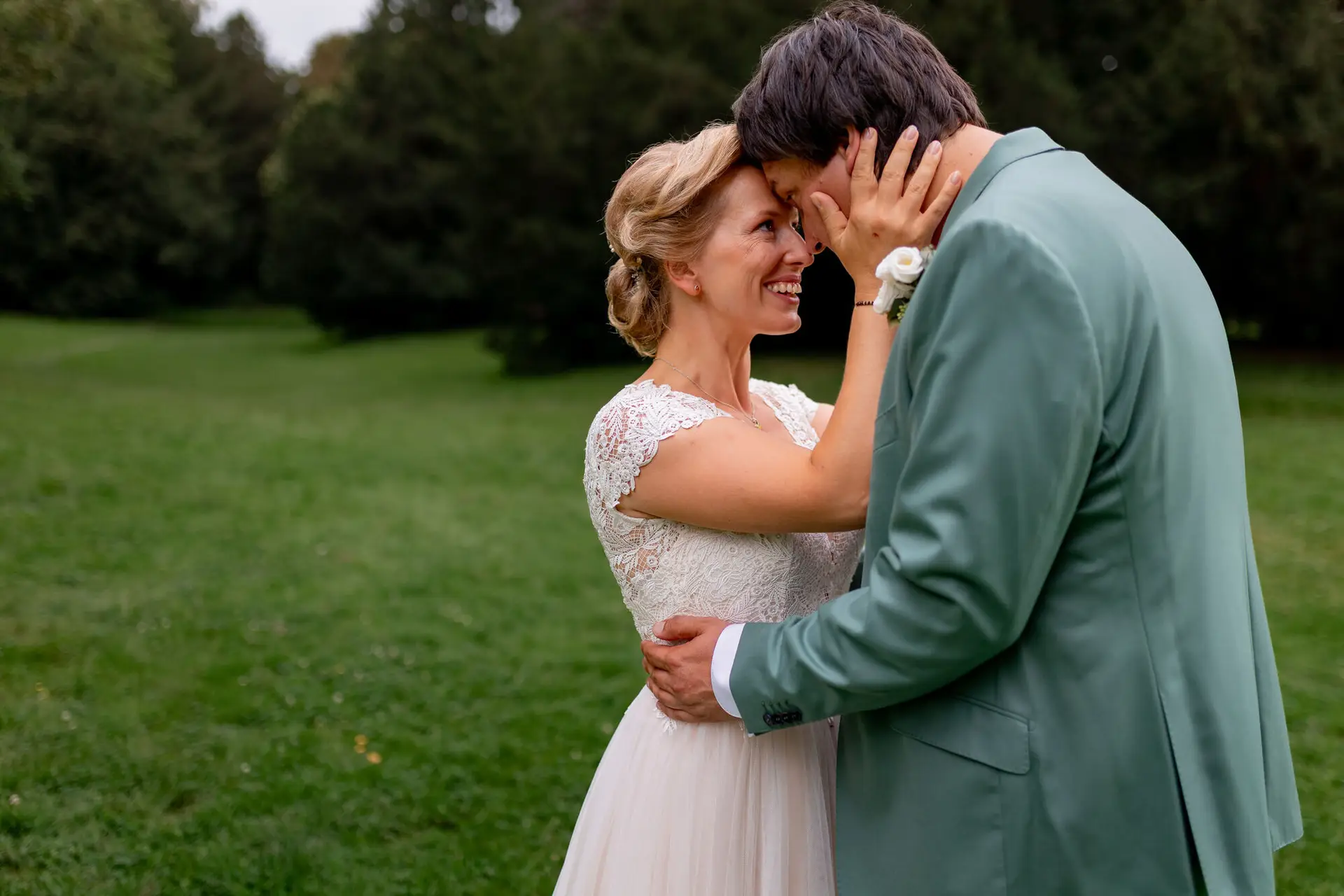 Strahlende Braut im weißen blumigen Kleid hält den Arm des Bräutigams, Braut lacht glücklich während sie das Gesicht des Bräutigams berührt im Schlossgarten Karlsruhe
