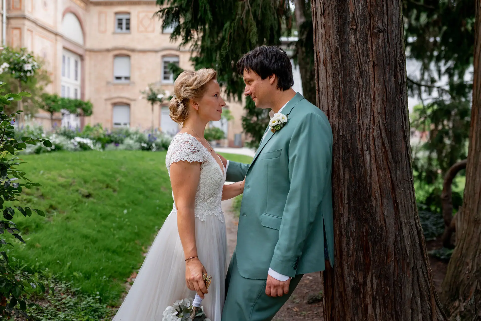 Brautpaar steht sich auf einem Gartenweg gegenüber und schaut sich liebevoll an, alter Baum und Gartenanlage im Hintergrund
