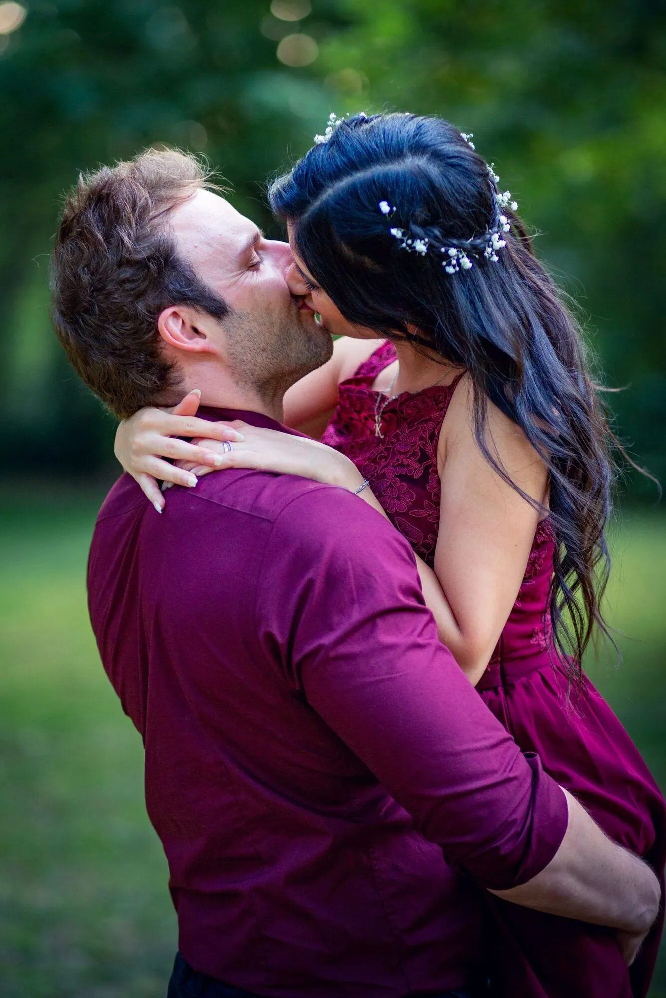 Davina und Keven küssen sich leidenschaftlich im Grünen, beide in bordeaux – sie im Spitzenkleid mit Blumenhaarschmuck, er im Hemd