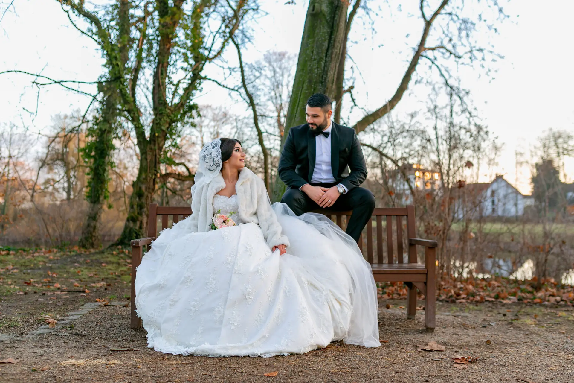 Brautpaar sitzt auf einer Holzbank im winterlichen Schlossgarten, Braut im weißen Fellmantel, kahle Bäume im Hintergrund