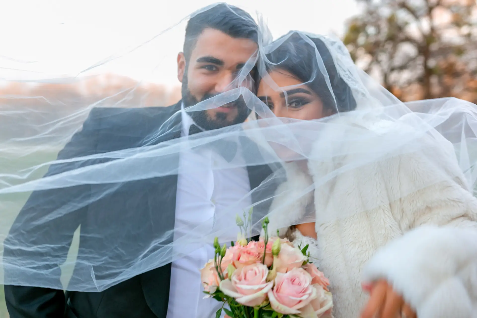 Brautpaar umschlingt sich innig unter weißem Schleier, rosa Blumenstrauß und kahle Äste im winterlichen Schlossgarten Worms-Herrnsheim