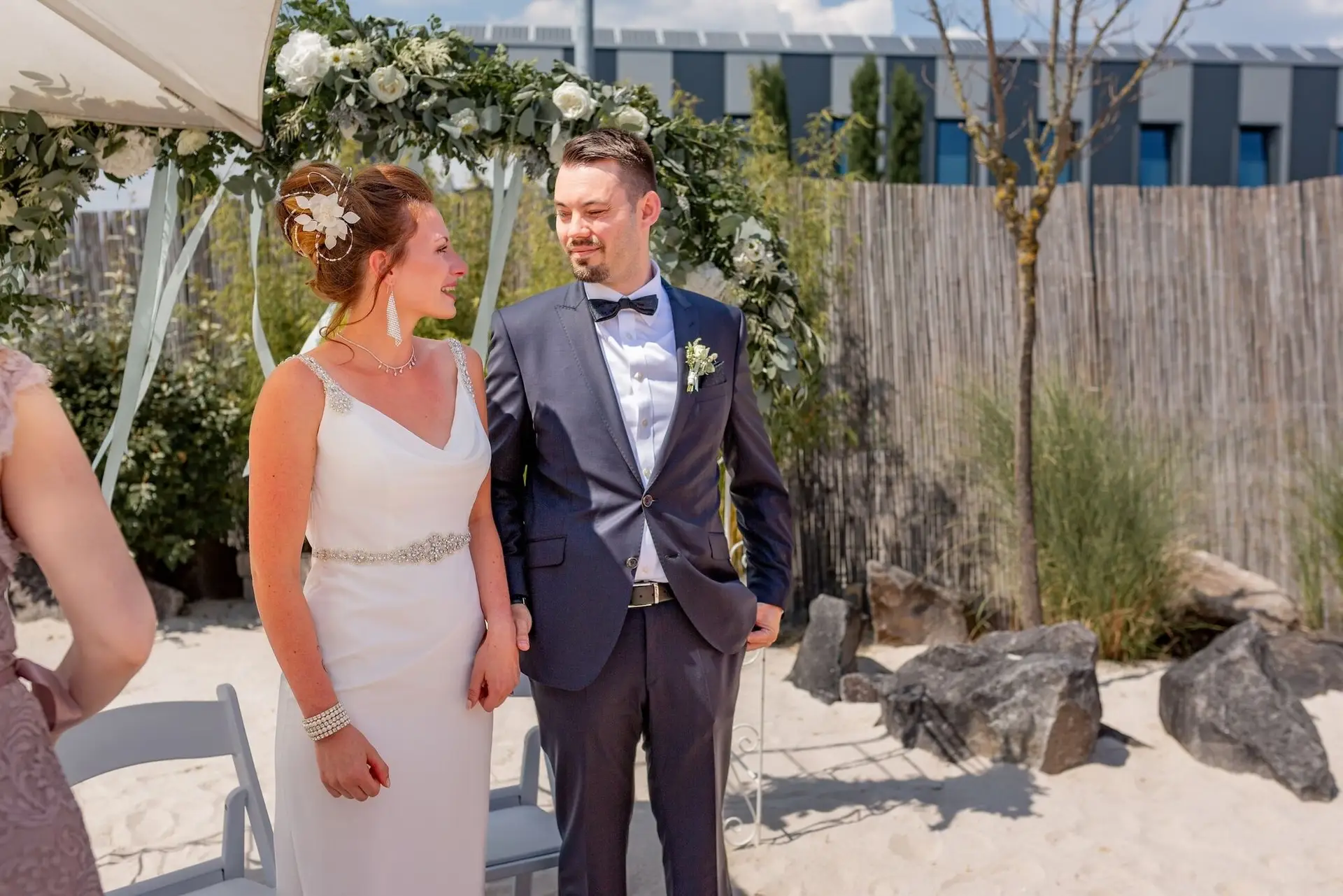 Brautpaar steht vereint unter dem weißen Blumenbogen, Sanduntergrund des Miio Schwetzingen