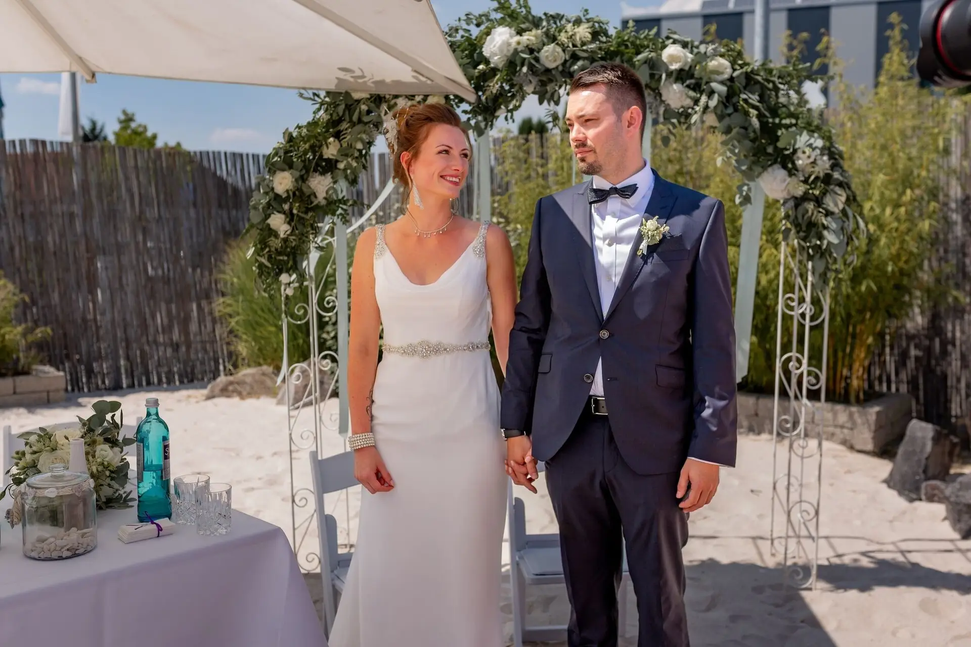 Glückliches Brautpaar küsst sich nach der Trauung vor dem weißen Blumenbogen, Sonnenstimmung