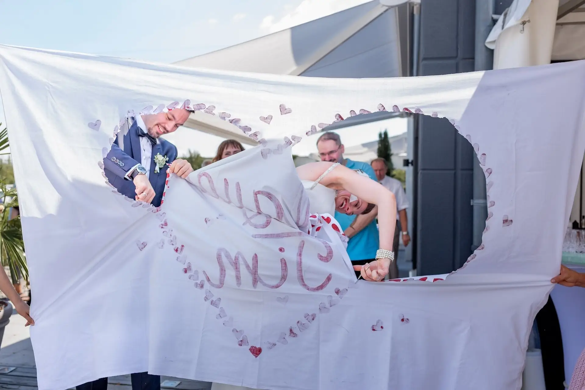 Bräutigam hält mit Braut ein großes "SMILE"-Tuch mit Herzchen, Gäste schauen lachend zu