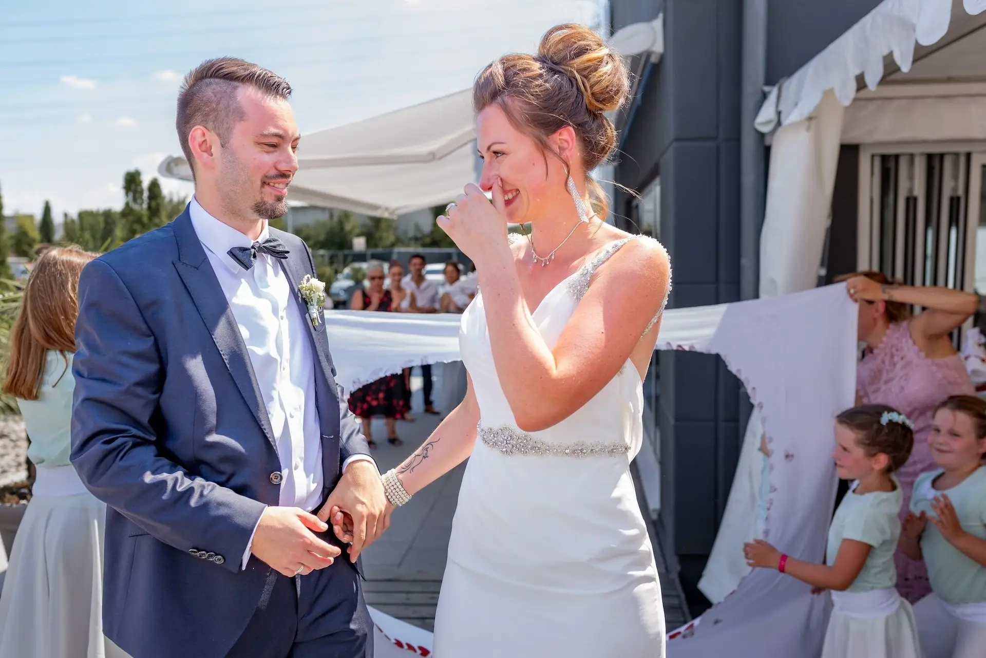 Bräutigam lächelt seiner gerührten Braut liebevoll an, Blumenmädchen und Gäste im Hintergrund