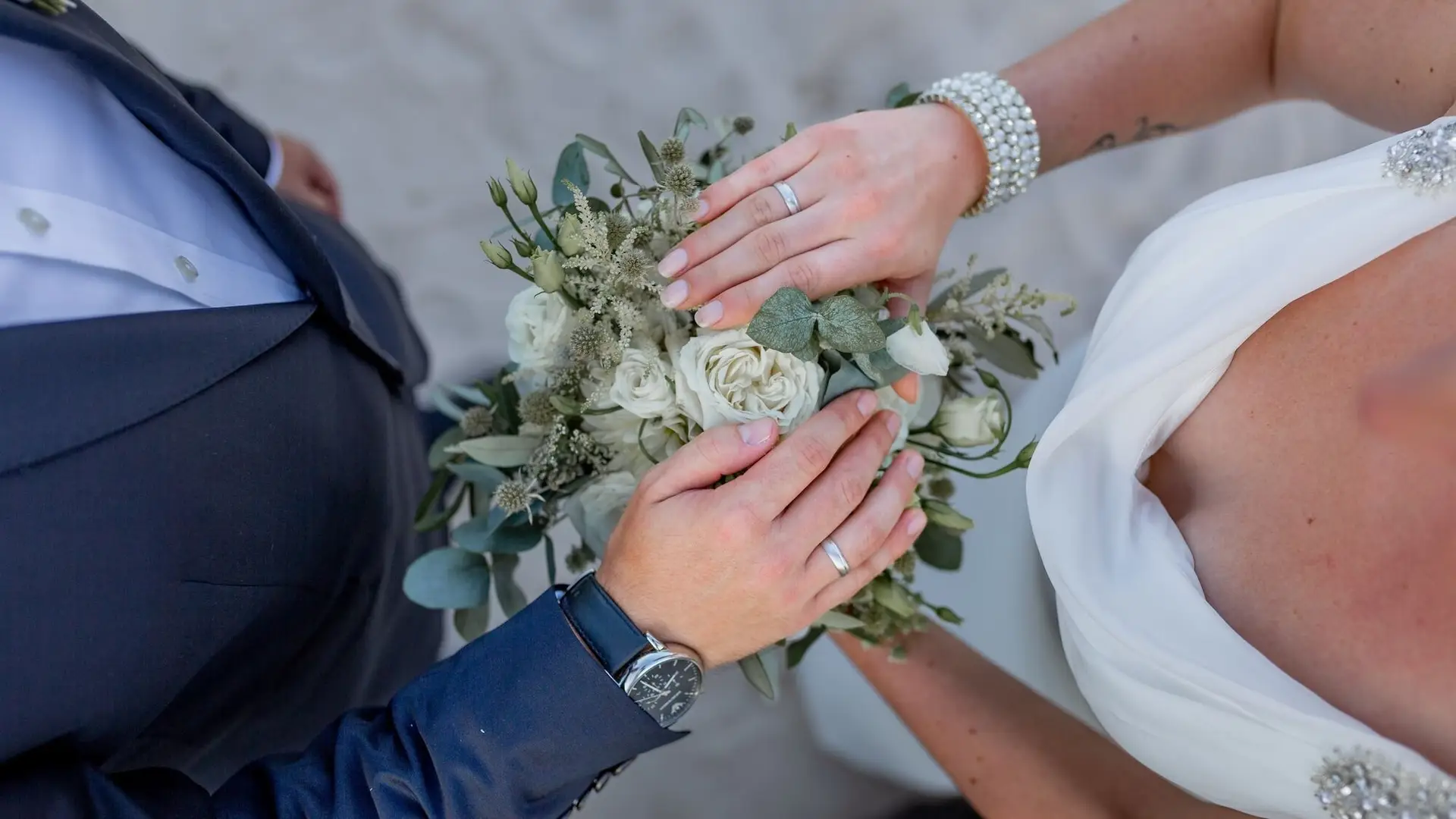 Nahaufnahme der verschränkten Hände des Brautpaares mit Blumenstrauß und den Eheringen