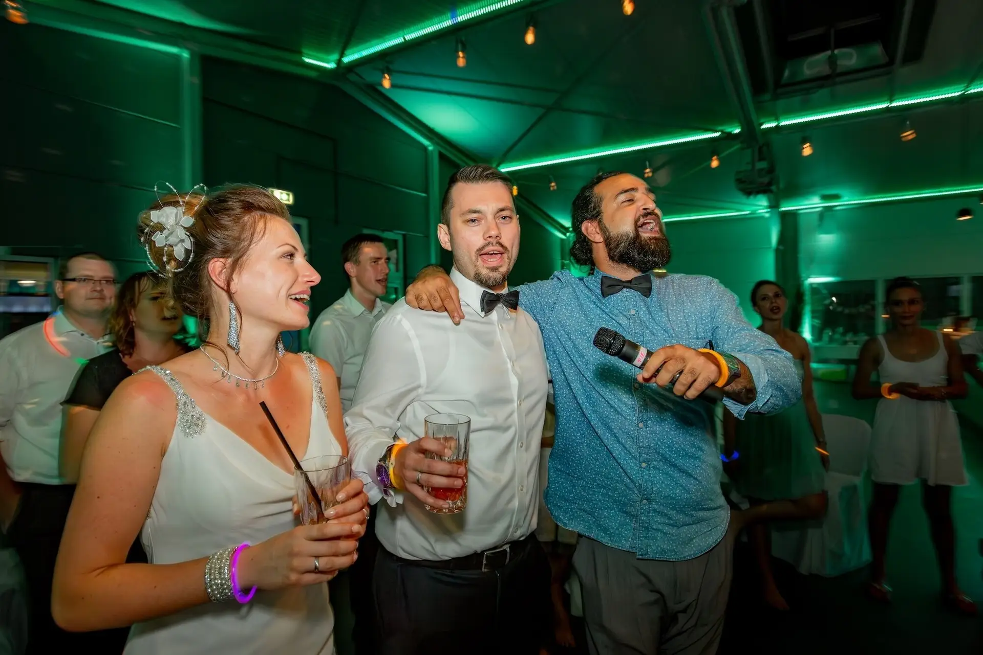 Bräutigam singt leidenschaftlich ins Mikrofon, umgeben von Braut und jubelnden Gästen bei grüner Party-Beleuchtung