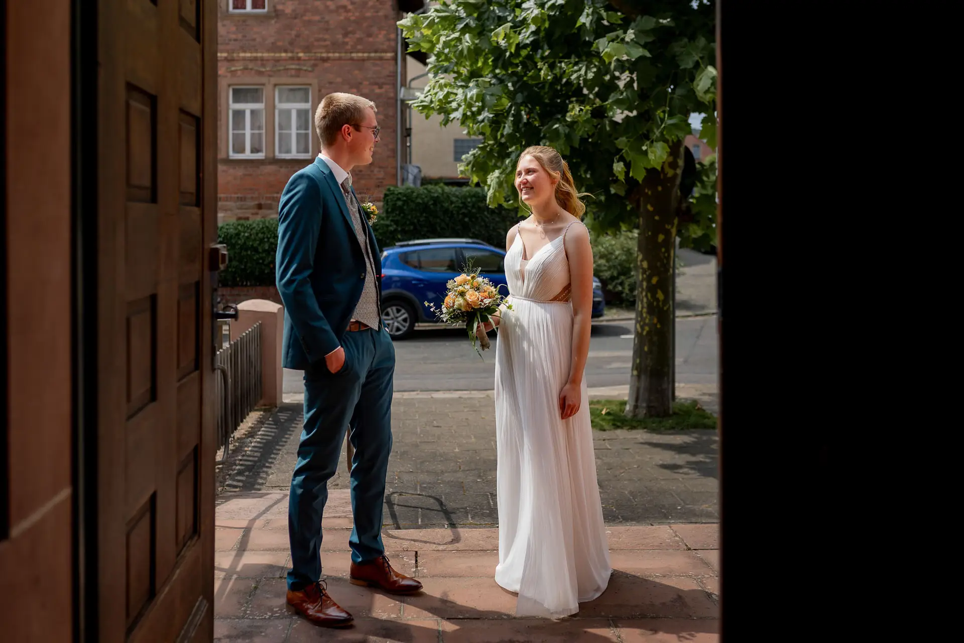 Brautpaar steht lächelnd vor der Kirche St. Johannes der Täufer in hellem Sonnenlicht
