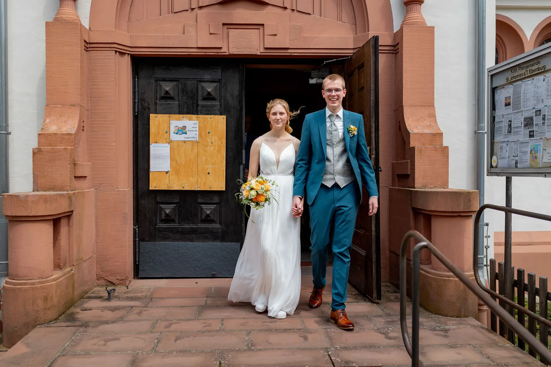 Brautpaar steht Hand in Hand vor dem Sandsteinportal der Kirche St. Johannes der Täufer