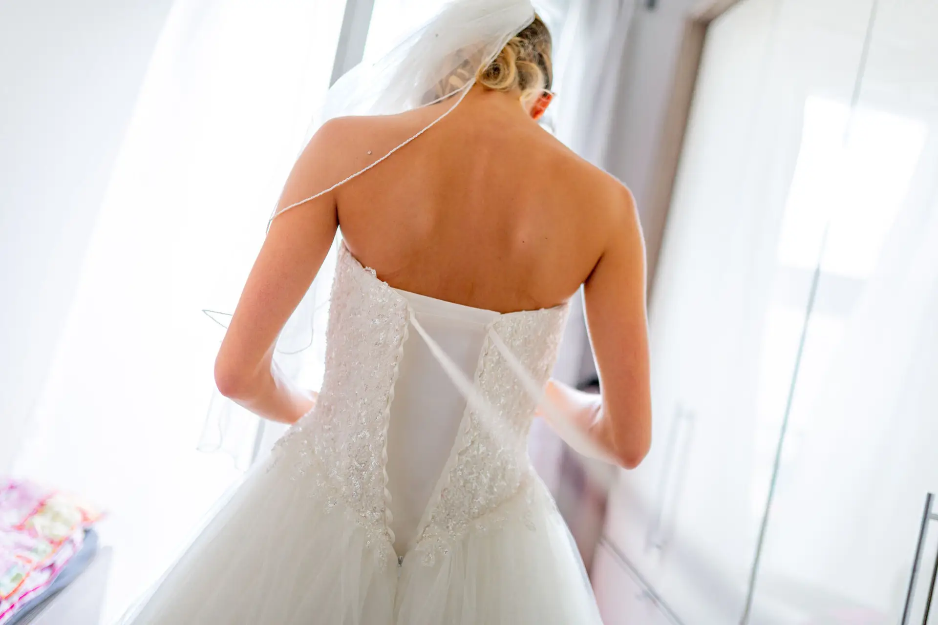Braut im Brautkleid am Fenster während Getting Ready, Rückansicht im Gegenlicht