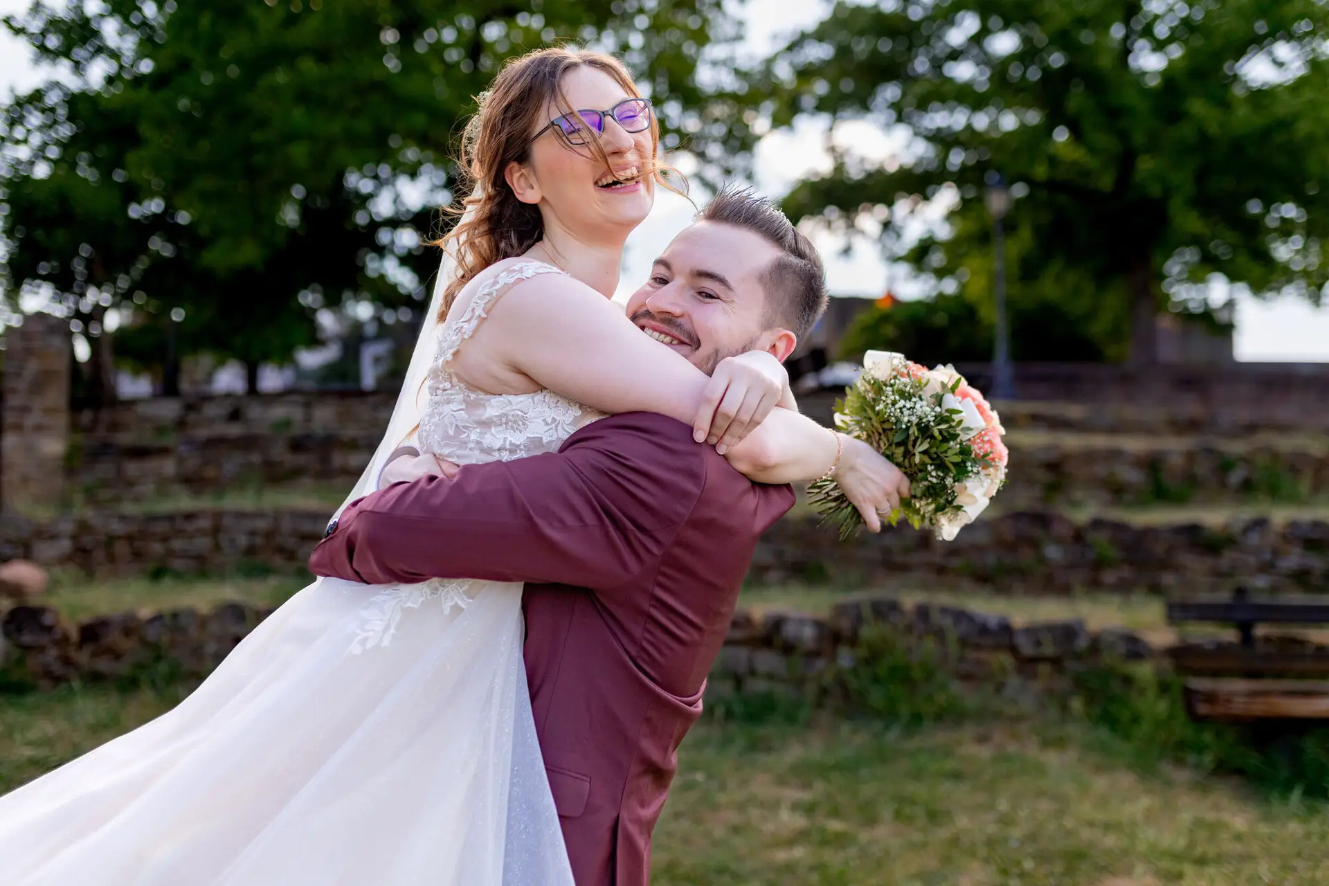 Bräutigam trägt seine lachende Braut ausgelassen hoch vor der Bruchsteinmauer der Burgschenke