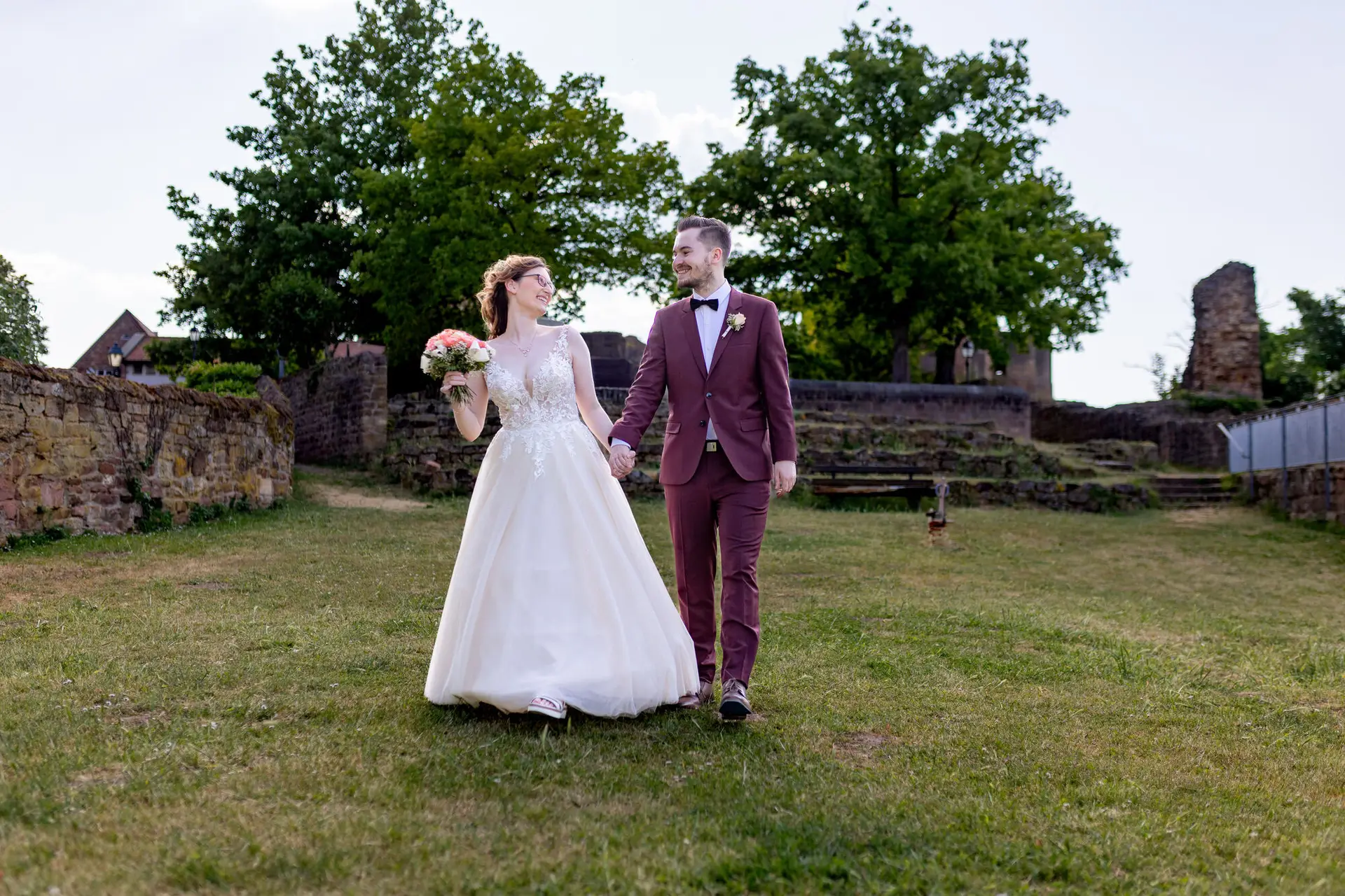 Brautpaar hält Händchen und spaziert auf dem Rasen entlang des historischen Steinwalls der Burganlage
