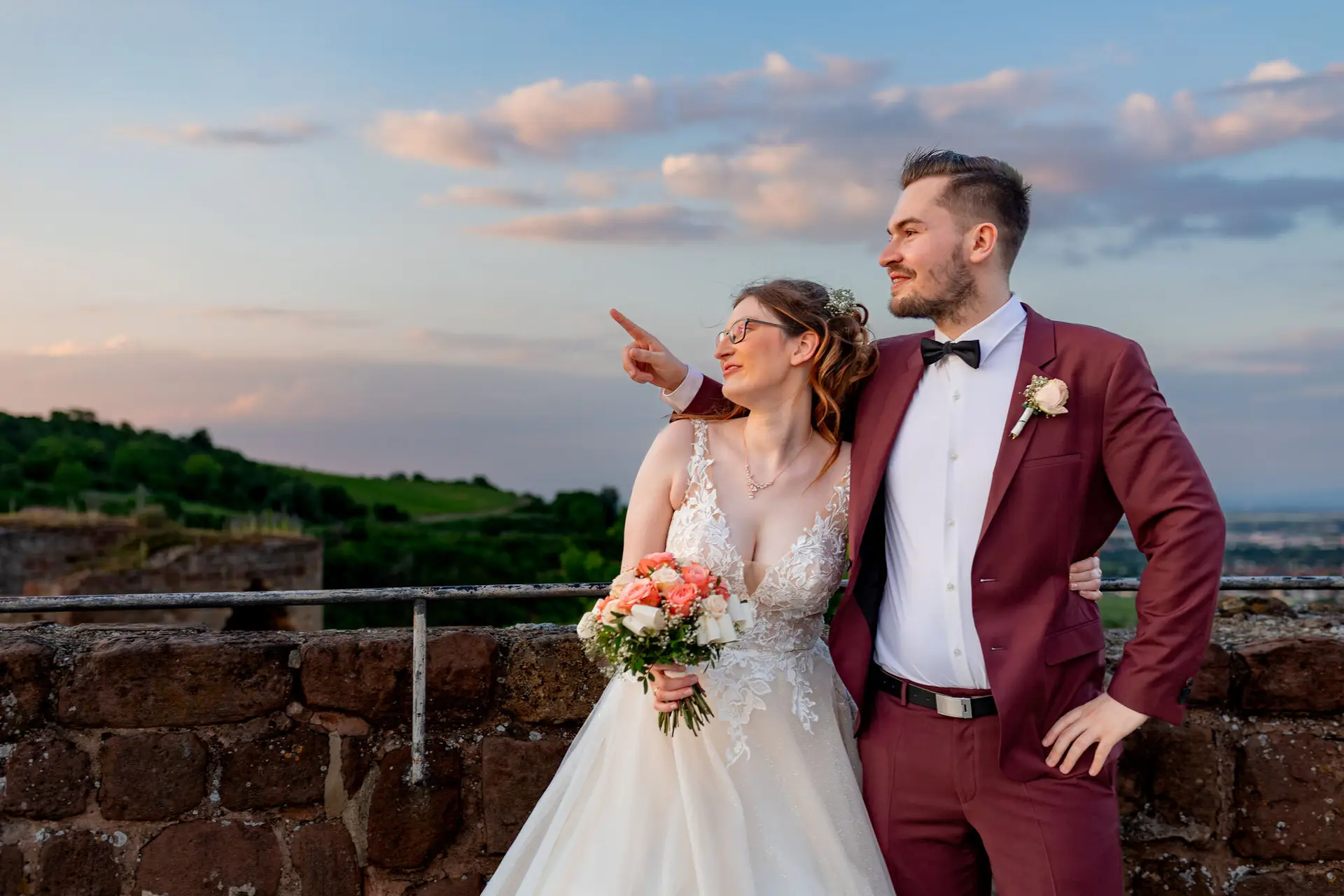 Brautpaar lacht herzlich und deutet auf den Horizont bei dramatischem Sonnenuntergang über der Burg Neuleiningen