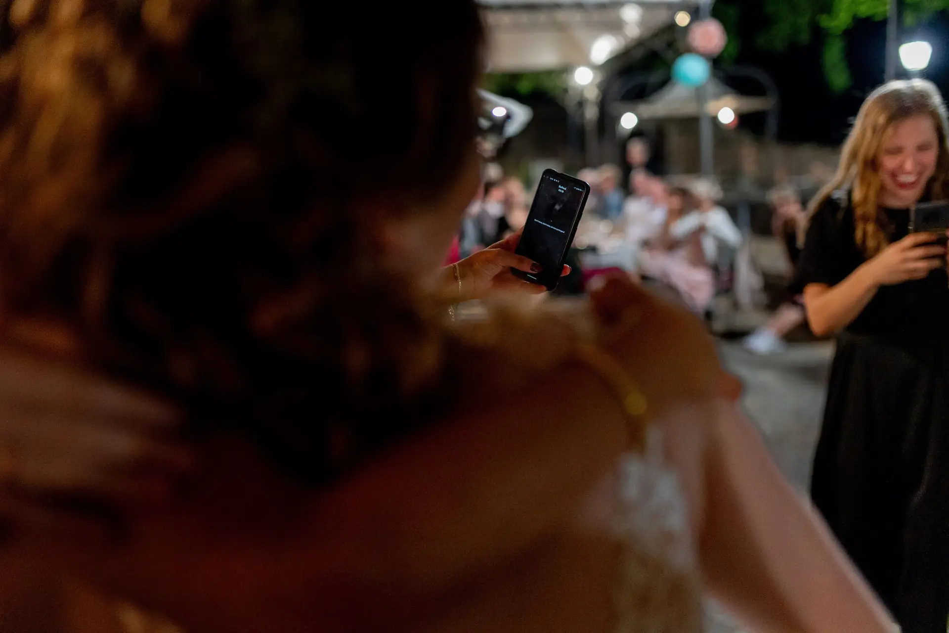 Braut hält Smartphone hoch und fotografiert feiernde Gäste in der Dämmerung bei der Hochzeitsparty