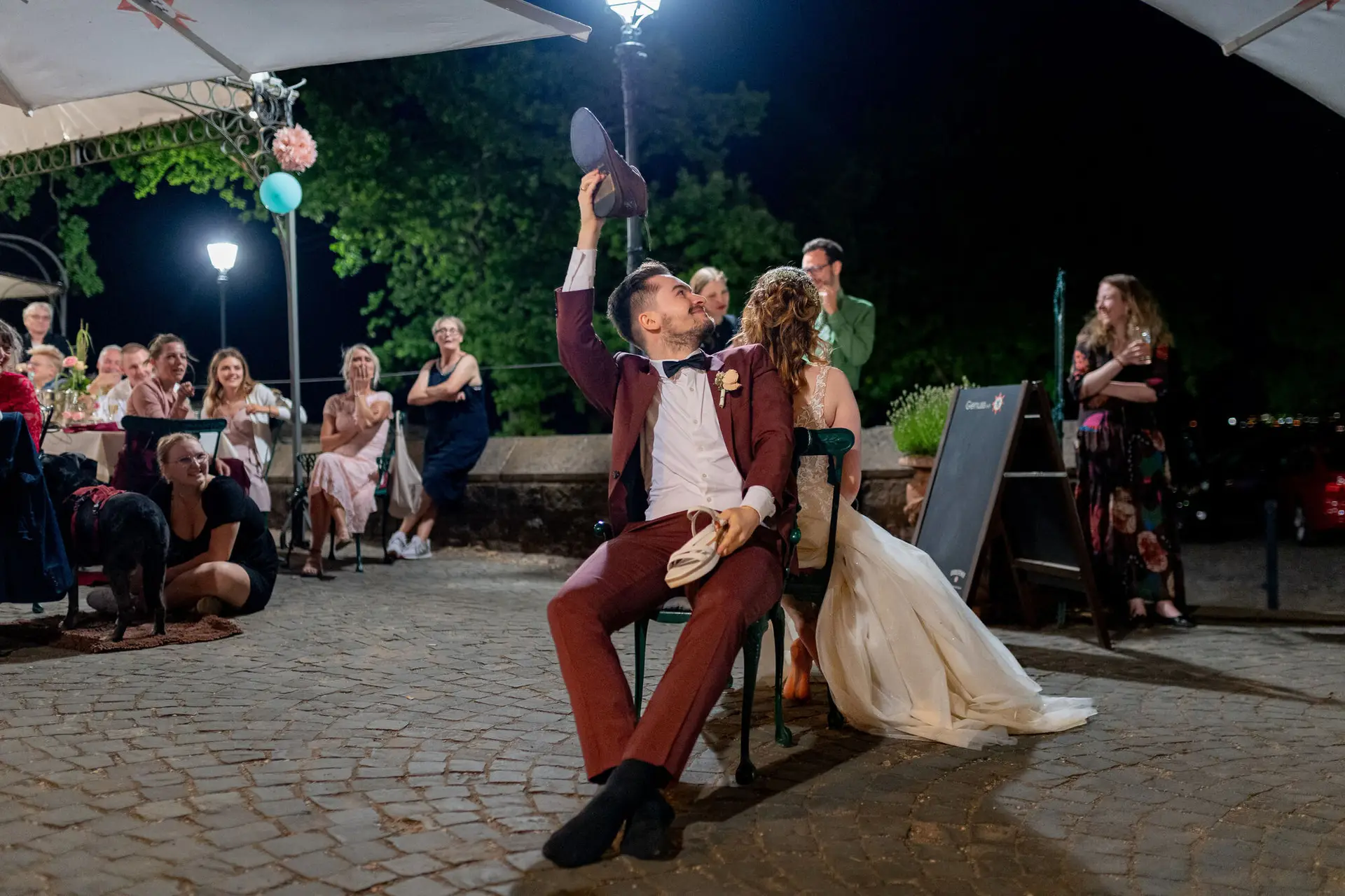 Schuhspiel auf der Hochzeitsfeier – Brautpaar sitzt Rücken an Rücken, Niklas hält einen Schuh hoch, Gäste schauen zu