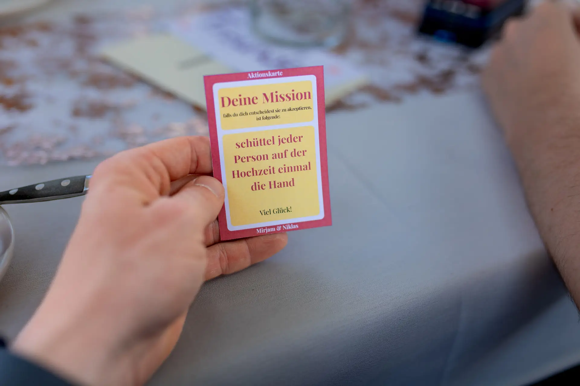 Hand hält eine rote Hochzeitskarte mit der Aufschrift „Viel Glück! Mirjam & Niklas" vor verschwommenem Hintergrund