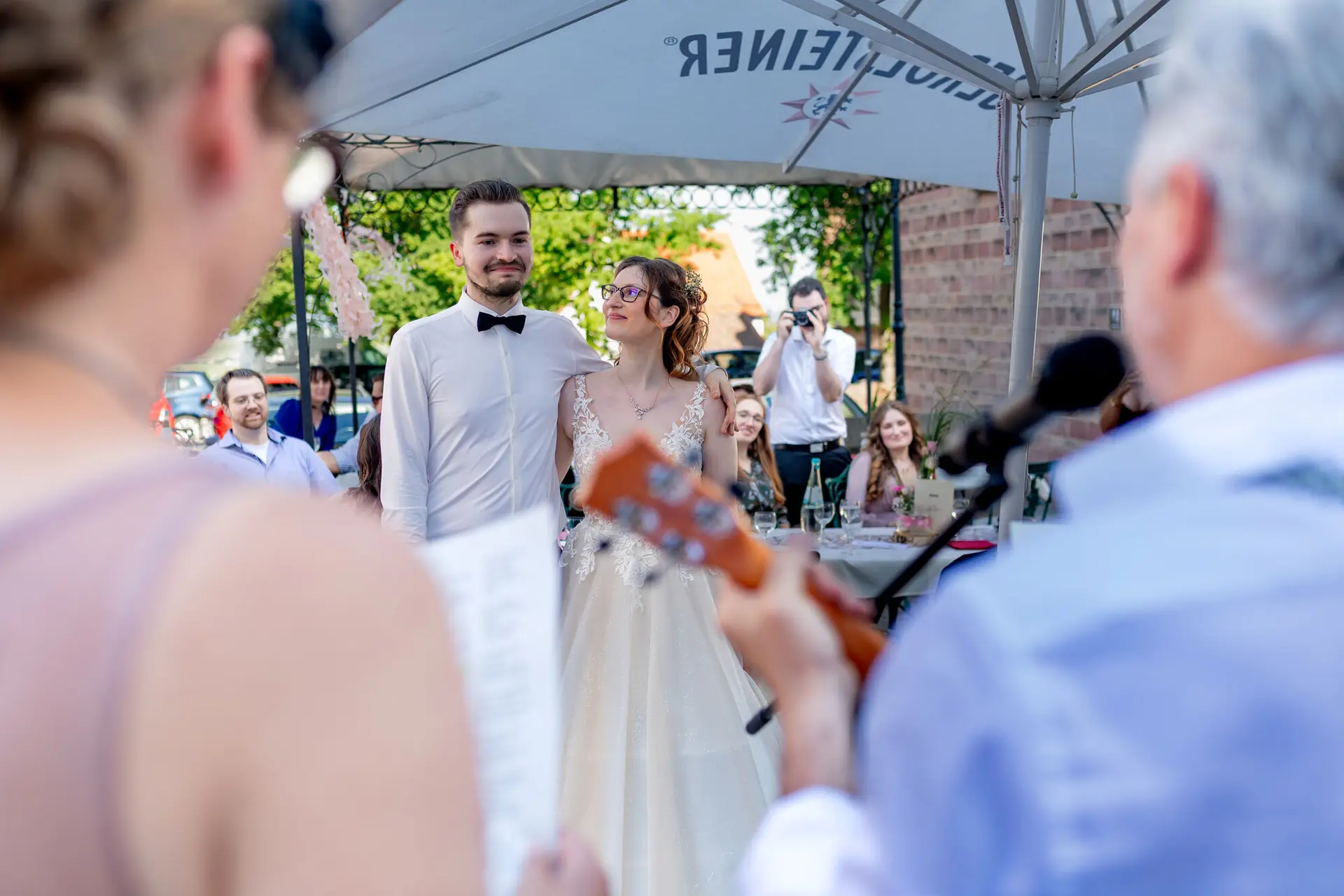 Brautpaar steht unter Sonnenschirm in Neuleiningen, Gast spielt Ukulele im Vordergrund