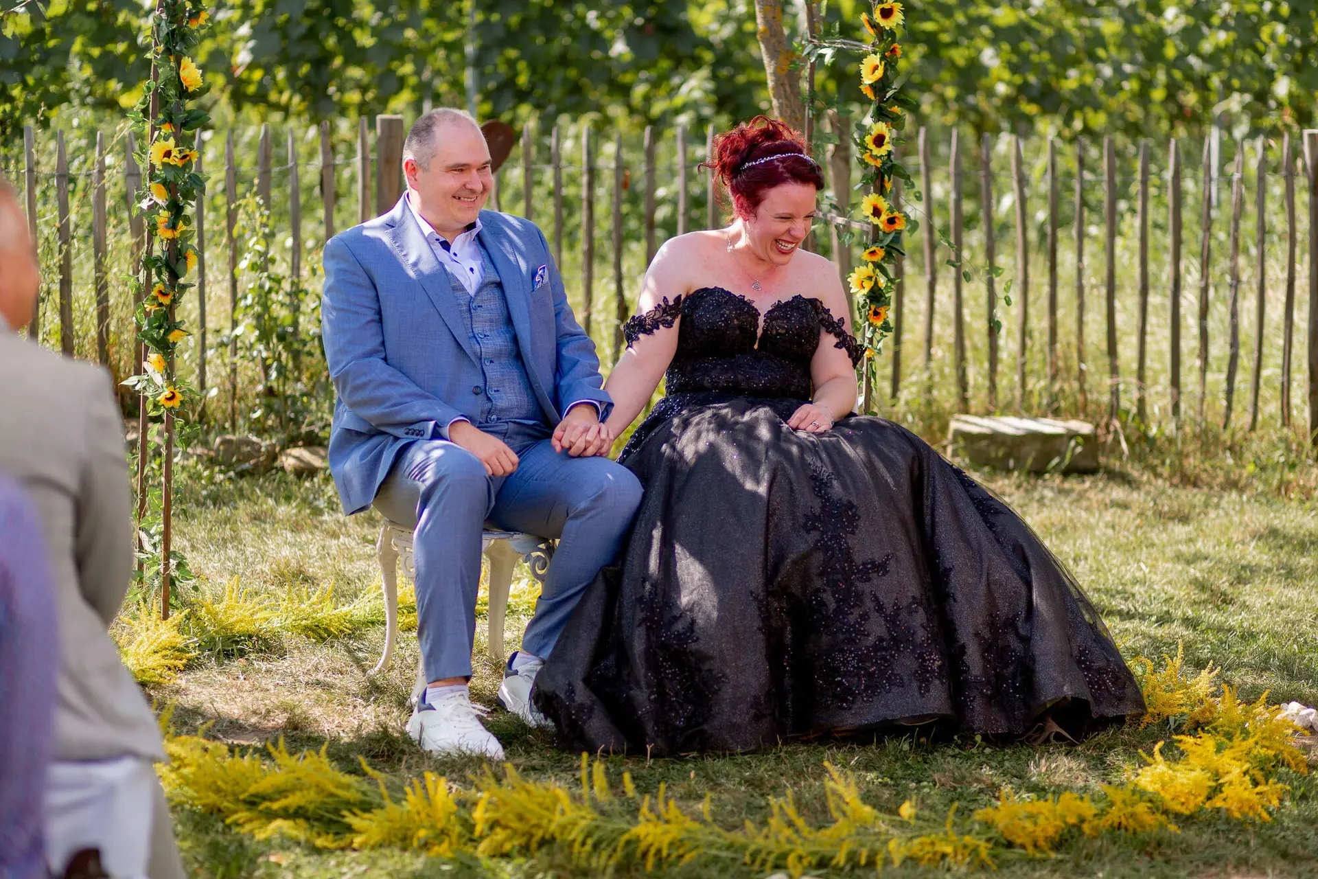 Brautpaar sitzt lachend unter Sonnenblumenbogen bei freier Trauung in Kellers Keller