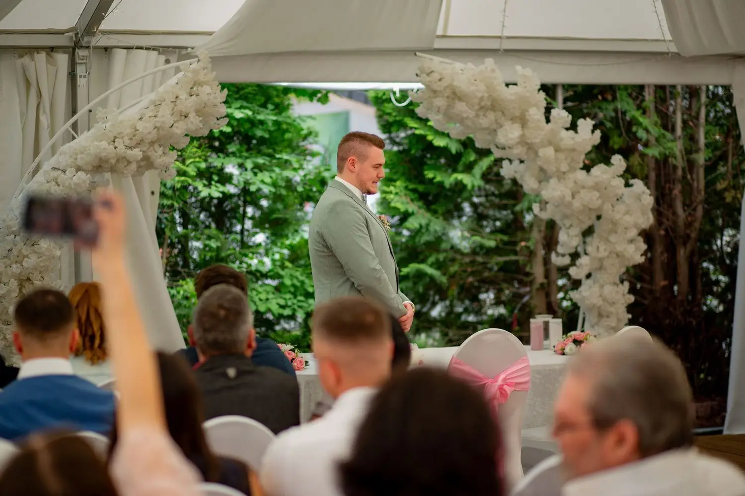Bräutigam wartet am weißen Blumenbogen beim Traualtar im Zelt, Hochzeitsgäste sitzen auf Stühlen dahinter