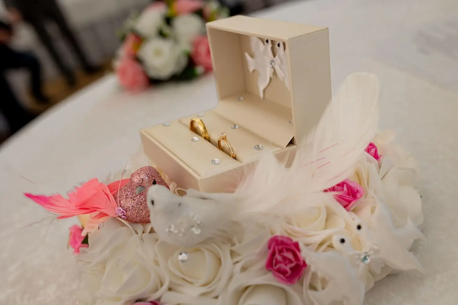 Zwei goldene Eheringe in beigem Ringkasten auf Blumentisch mit rosafarbenen Blüten