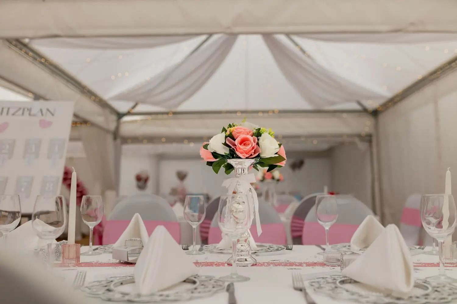 Elegant gedeckter Tisch im weißen Festzelt mit rosa Schleifen an den Stühlen und Blumenarrangement