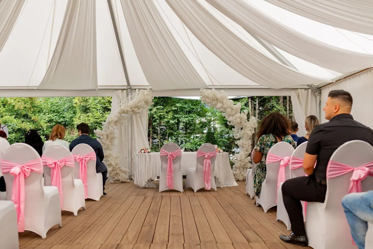Hochzeitsgäste sitzen unter weißem Zelt bei der freien Trauung, weißer Blumenbogen als Altar, Bäume im Hintergrund