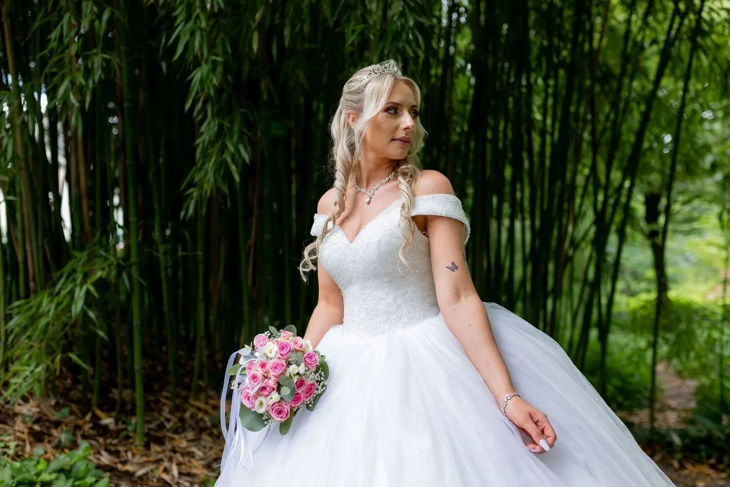 Braut in glitzerndem weißem Ballkleid mit Blumenstrauß vor dichten Bambus-Pflanzen des Hermannshof-Gartens