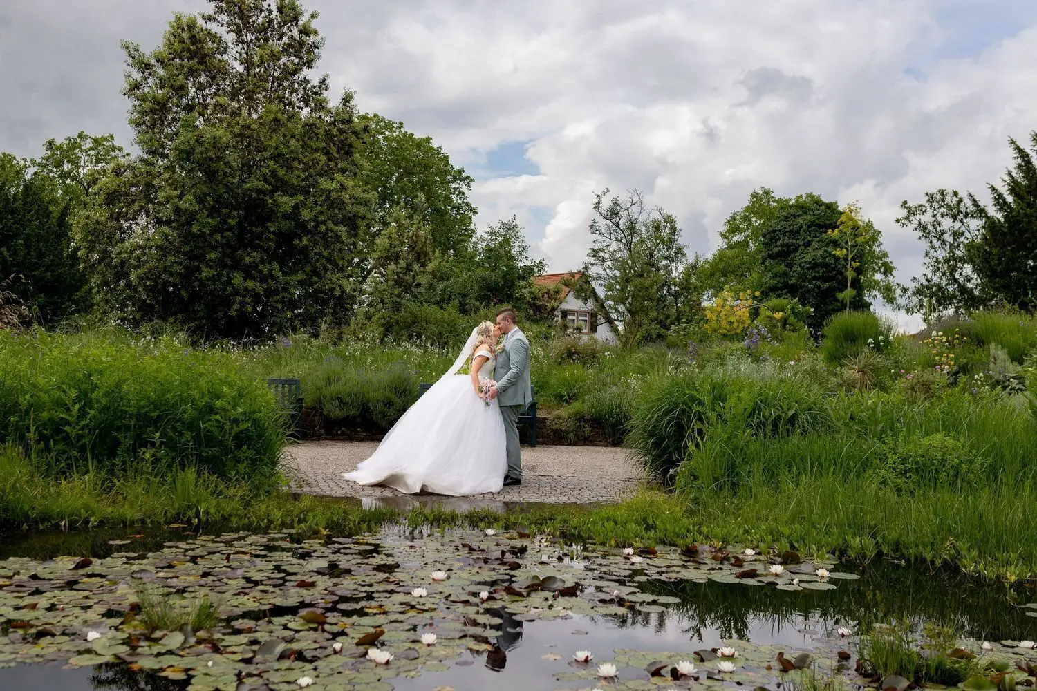 Brautpaar küsst sich innig am Teich mit Seerosenblättern, üppige Bäume und Garten im Hintergrund