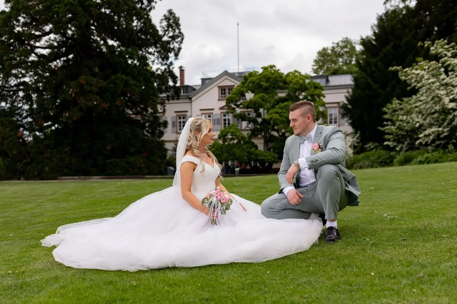 Brautpaar sitzt auf der Wiese und unterhält sich, historisches Herrenhaus des Hermannshofs im Hintergrund