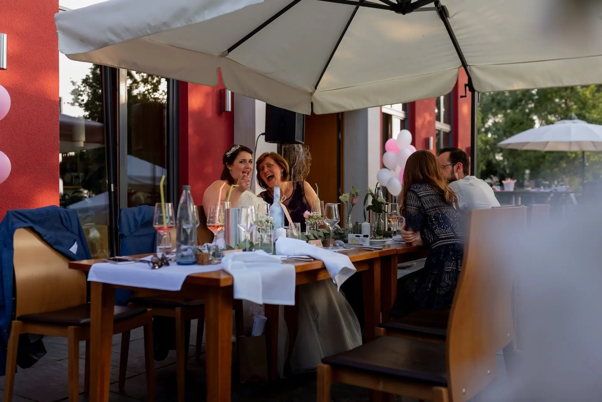 Hochzeitsgesellschaft am Tisch im Freien mit Rosengestecken, pinken Luftballons und weißen Tischdecken