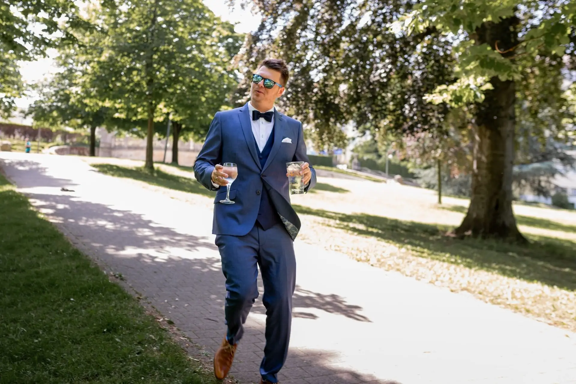 Bräutigam im blauen Anzug schlendert entspannt mit zwei Gläsern durch den Kurpark Bad Dürkheim