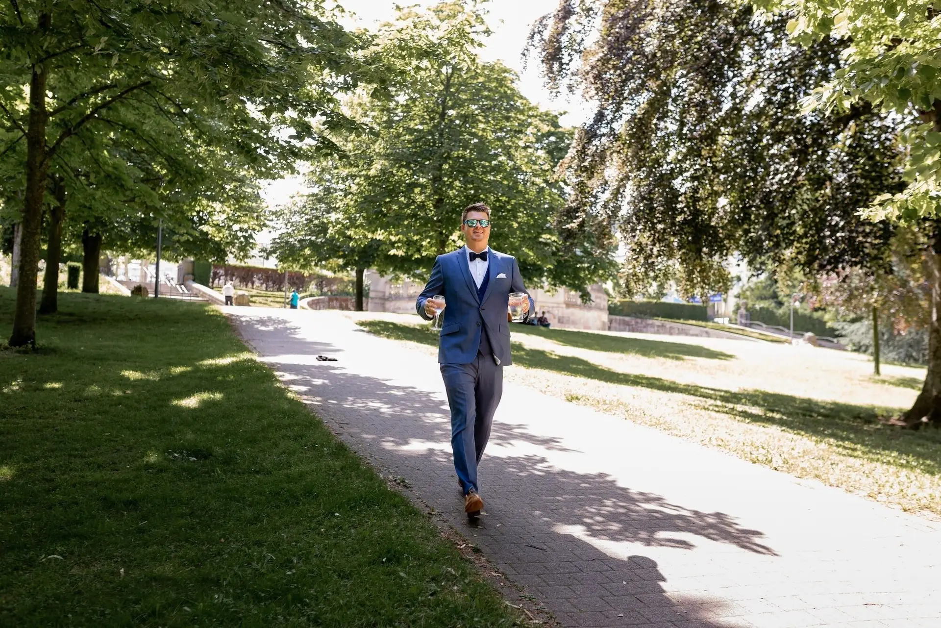 Bräutigam im dunkelblauen Anzug und Sonnenbrille bringt zwei Gläser durch den Kurpark Bad Dürkheim