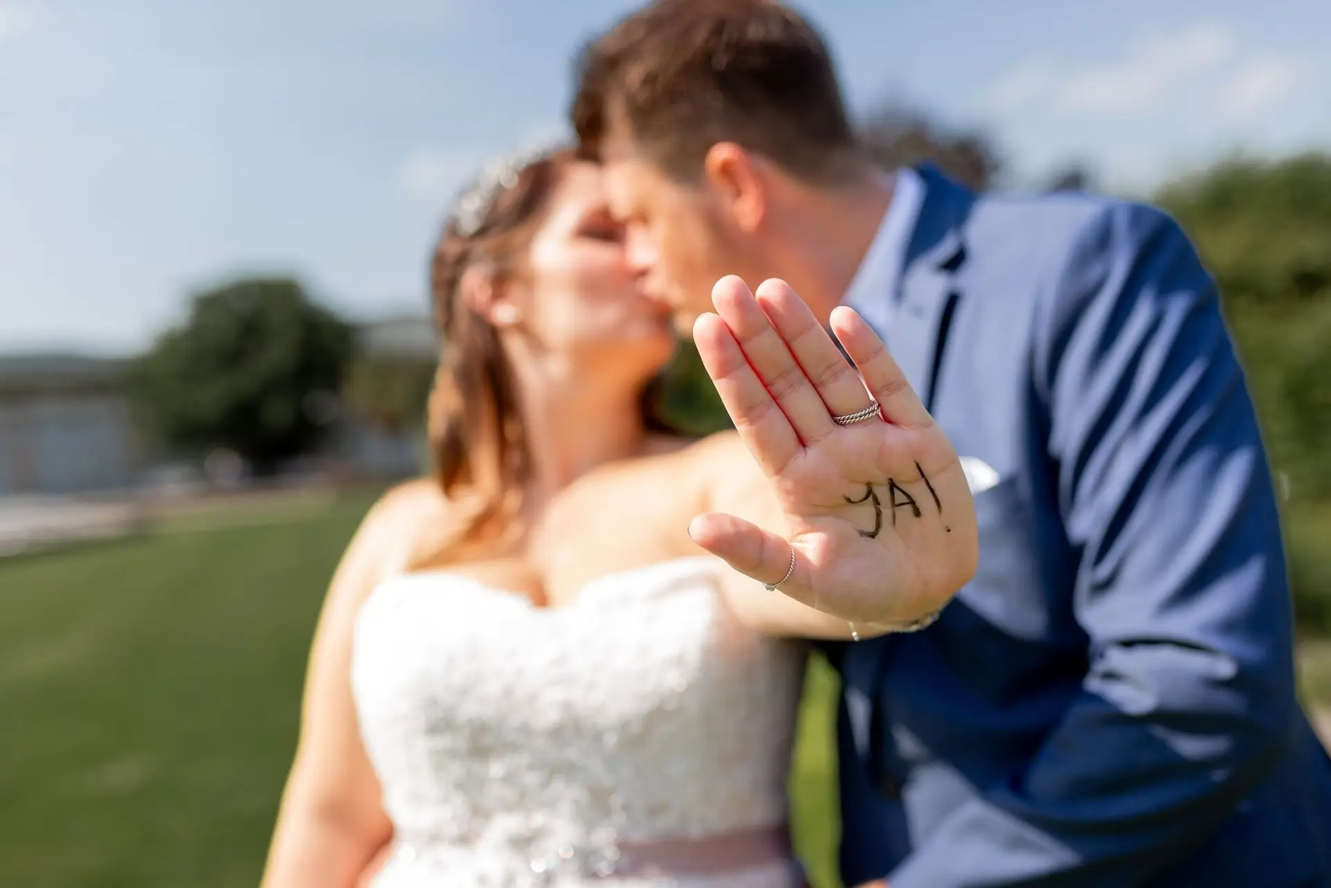 Brautpaar küsst sich, Bräutigam zeigt Hand mit aufgeschriebenem "JA" im Kurpark Bad Dürkheim