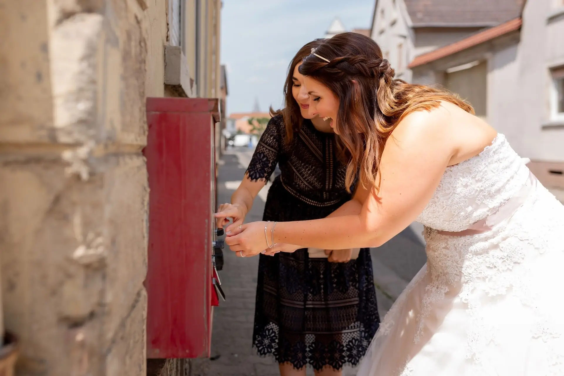 Braut und Freundin spielen lachend an einem roten Kaugummiautomaten in der Gasse von Mutterstadt